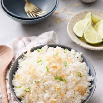 Pinterest graphic of a bowl of coconut rice topped with green onions and toasted coconut. Bowl of limes in the back along with more garnishes and bowls with forks.