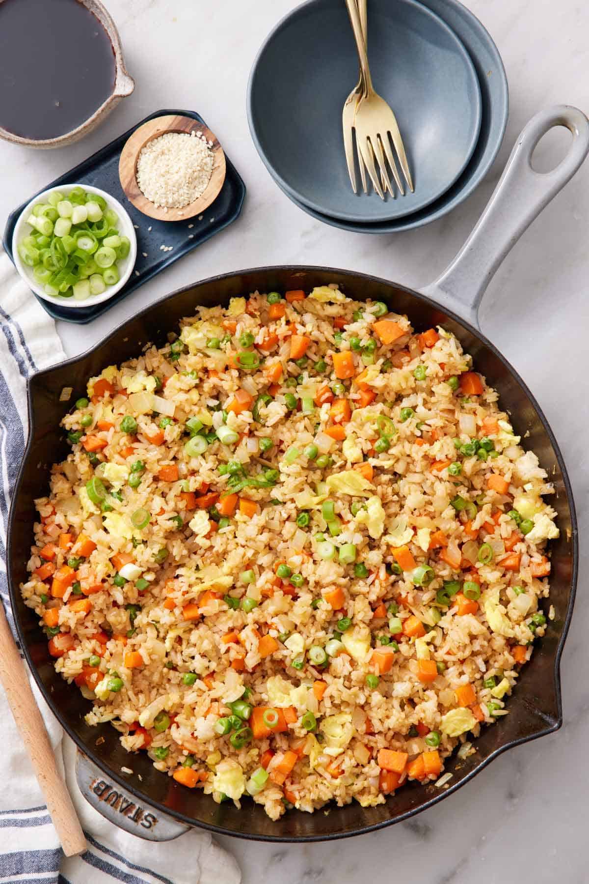 An overhead view of a skillet of fried rice with a stack of bowls, forks, and bowls of garnishes on the side.