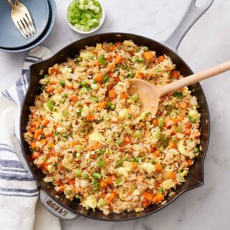 Overhead view of a skillet of fried rice with a wooden spoon tucked inside. Green onions, bowls, and forks on the side.