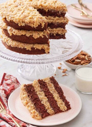 A slice of German chocolate cake on a plate in front of a cake stand with the rest of the cake.