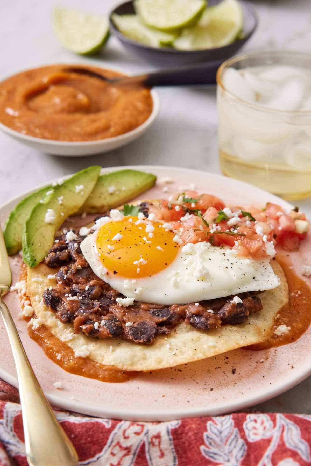 A profile view of huevos rancheros on a plate, showing the layers of sauce, tortilla, and toppings. A drink, sauce, and limes in the background.