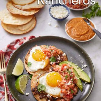 Pinterest graphic of a plate with a serving of huevos rancheros along with lime wedges and avocado slices. Additional toppings in bowls and more tortillas in the background.