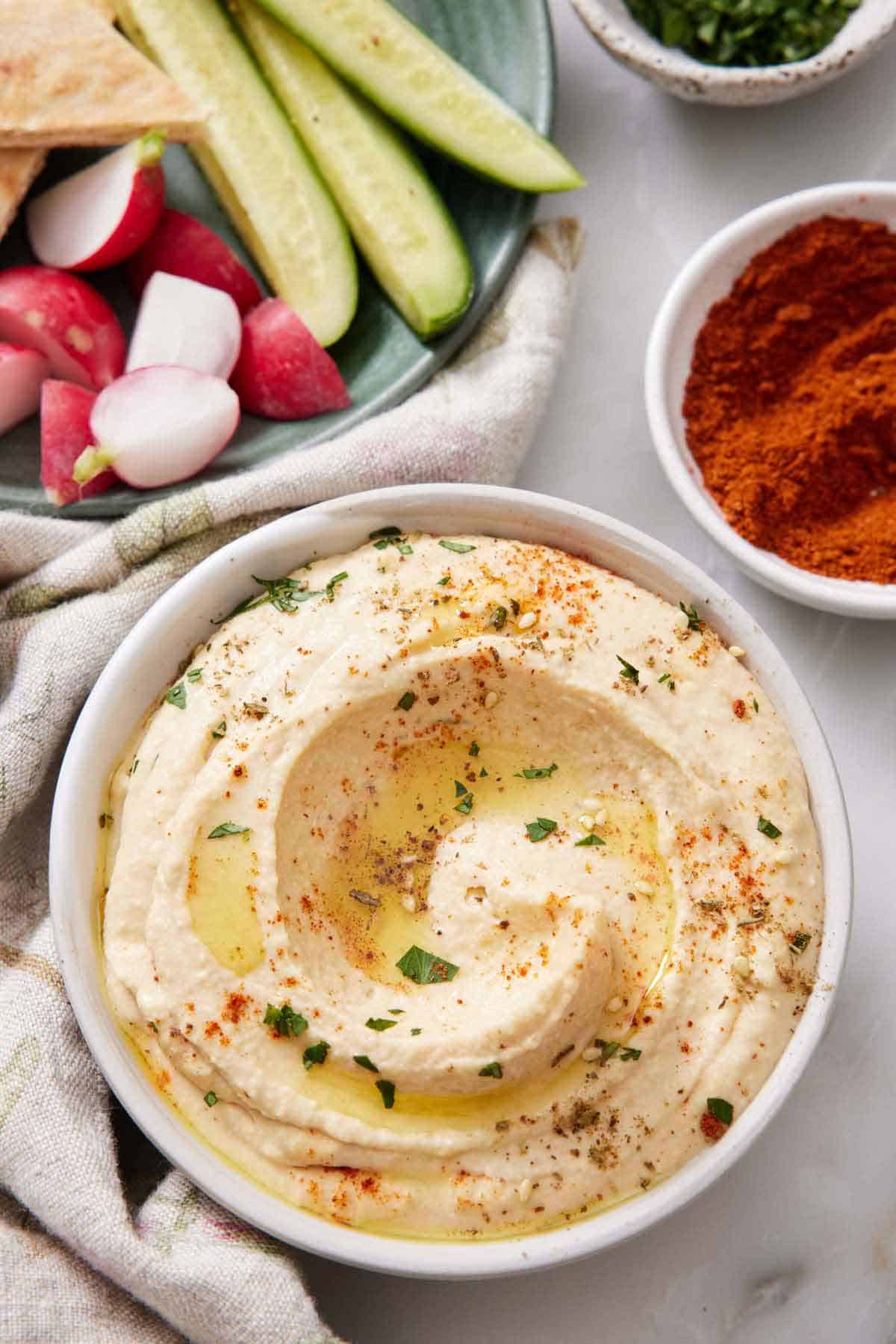 Overhead view of a bowl of hummus with oil, parsley, and paprika as garnish. A bowl of cucumbers and radish in the back with a bowl of paprika on the side.