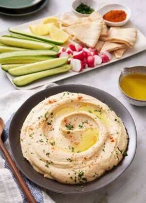 A bowl of hummus topped with oil, paprika, and parsley. Cucumbers, radish, lemons, pita bread, and garnishes in the background on a platter.