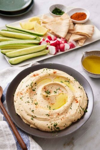 A bowl of hummus topped with oil, paprika, and parsley. Cucumbers, radish, lemons, pita bread, and garnishes in the background on a platter.