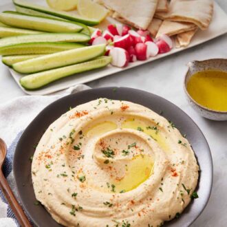 Pintrest graphic of a bowl of hummus topped with oil, paprika, and parsley with a platter of dippers in the background.