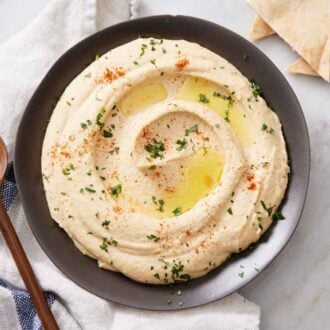 Overhead view of a bowl of hummus with olive oil, chopped parsley, and paprika garnished on top. A spoon and some pita bread on the side.