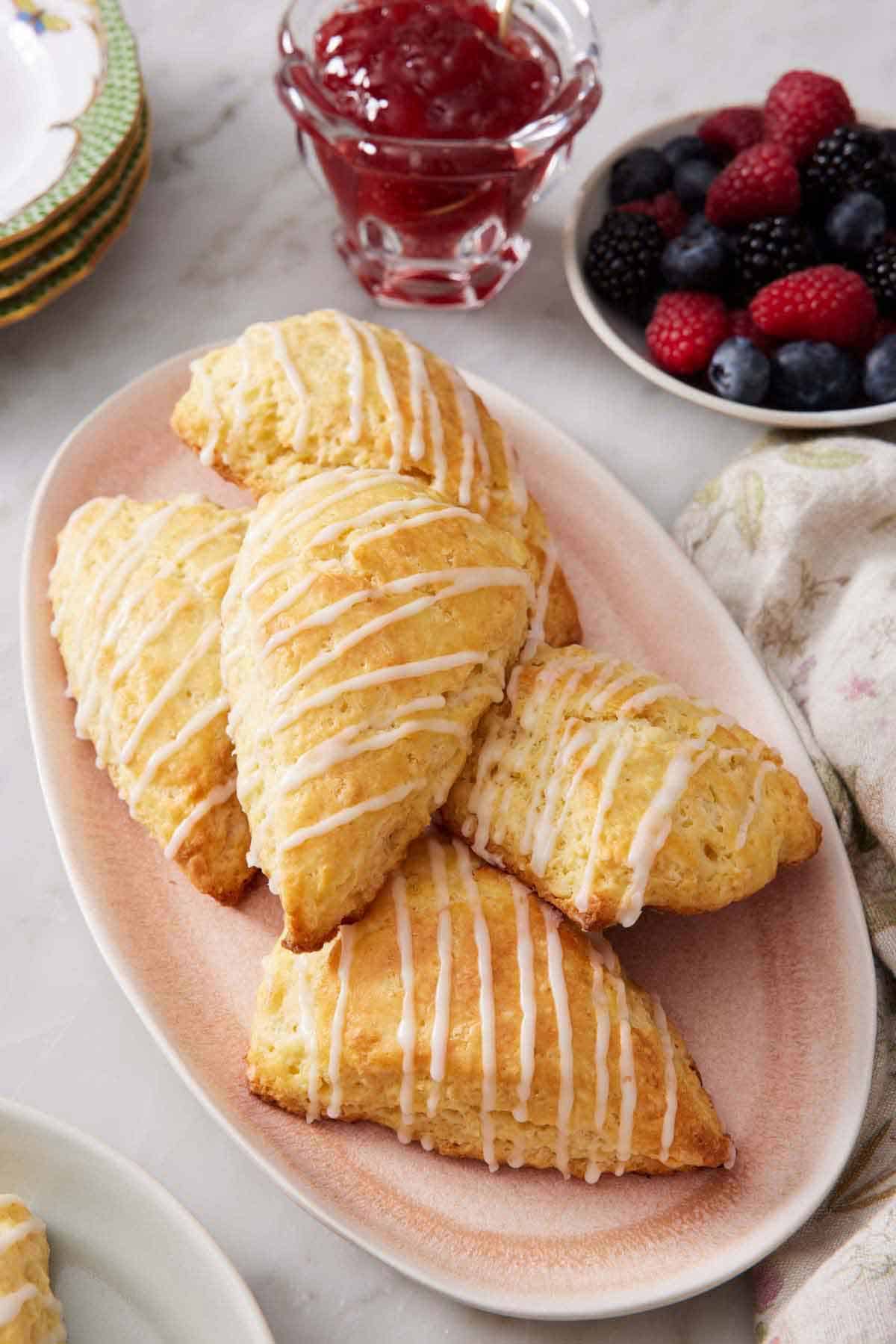 A pink platter of lemon scones with a bowl of berries and a bowl of jam in the back.