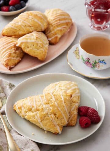 A plate with two lemon scones with fresh raspberries. A cup of tea and additional scones in the background on a platter.