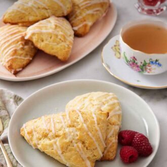 Pinterest graphic of a plate with two lemon scones with fresh raspberries. A cup of tea and additional scones in the background on a platter.