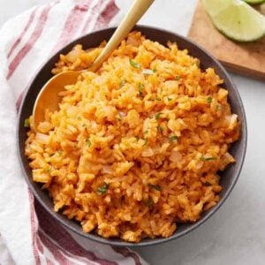 Overhead view of a bowl of Mexican rice with a spoon inside. Cut limes and a napkin on the side.