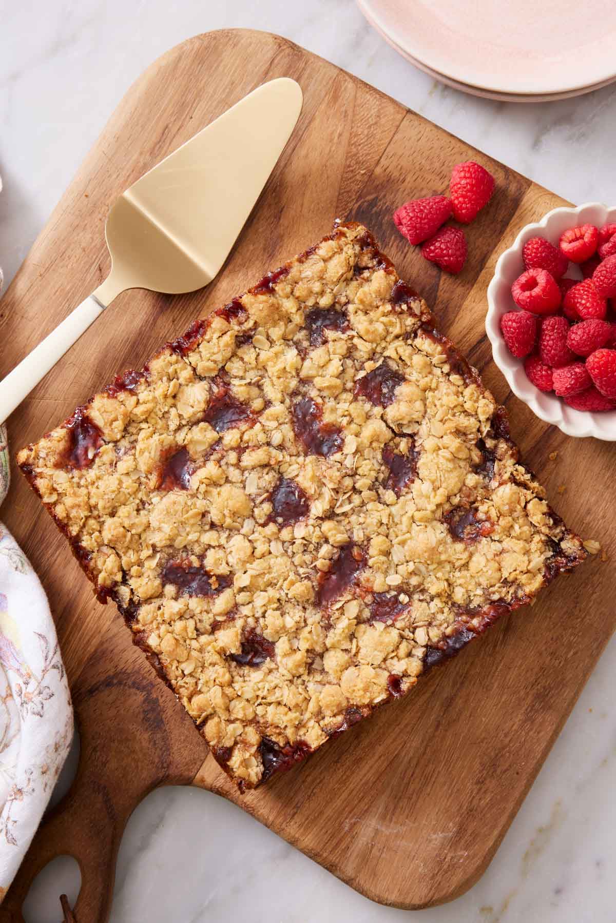 Overhead view of a slab of uncut raspberry bars on a wooden serving board with fresh raspberries on the side.
