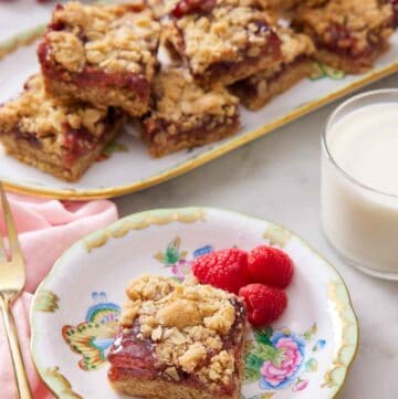 A plate with a piece of raspberry bars with three fresh raspberries. A glass of milk and a platter of more bars in the background.