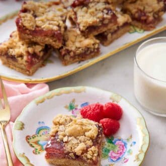 Pinterest graphic of a plate with a piece of raspberry bars with three fresh raspberries. A glass of milk and a platter of more bars in the background.