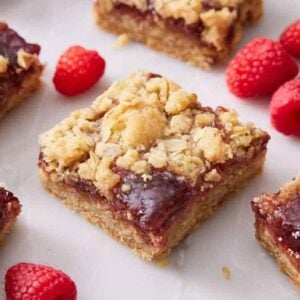 Multiple pieces of raspberry bars on a marble surface with fresh raspberries scattered around.