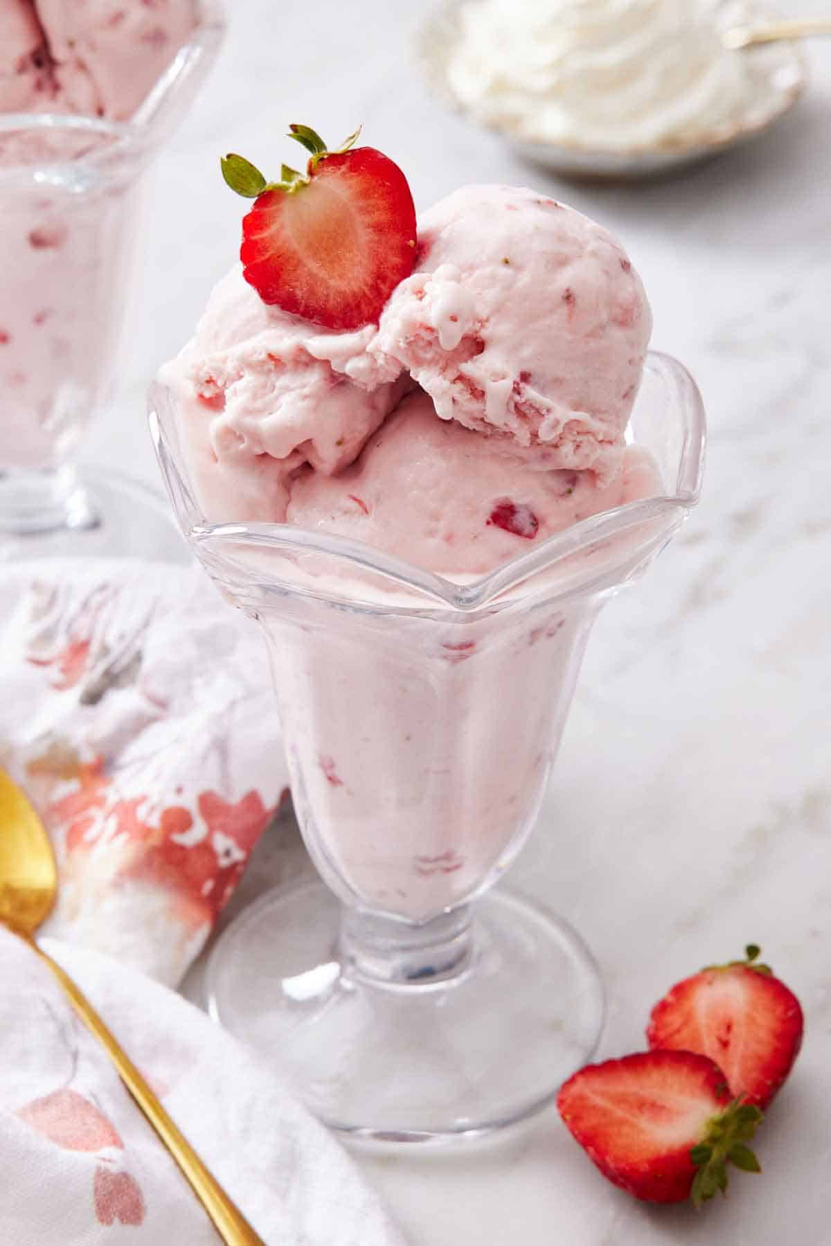 A sundae glass with strawberry ice cream topped with cut a strawberry. More strawberries beside it.