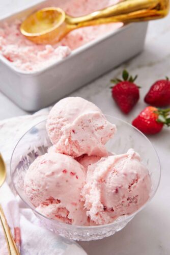 A bowl with scoops of strawberry ice cream with fresh strawberries in the background along with a container of more ice cream.