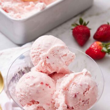A bowl with scoops of strawberry ice cream with fresh strawberries in the background along with a container of more ice cream.