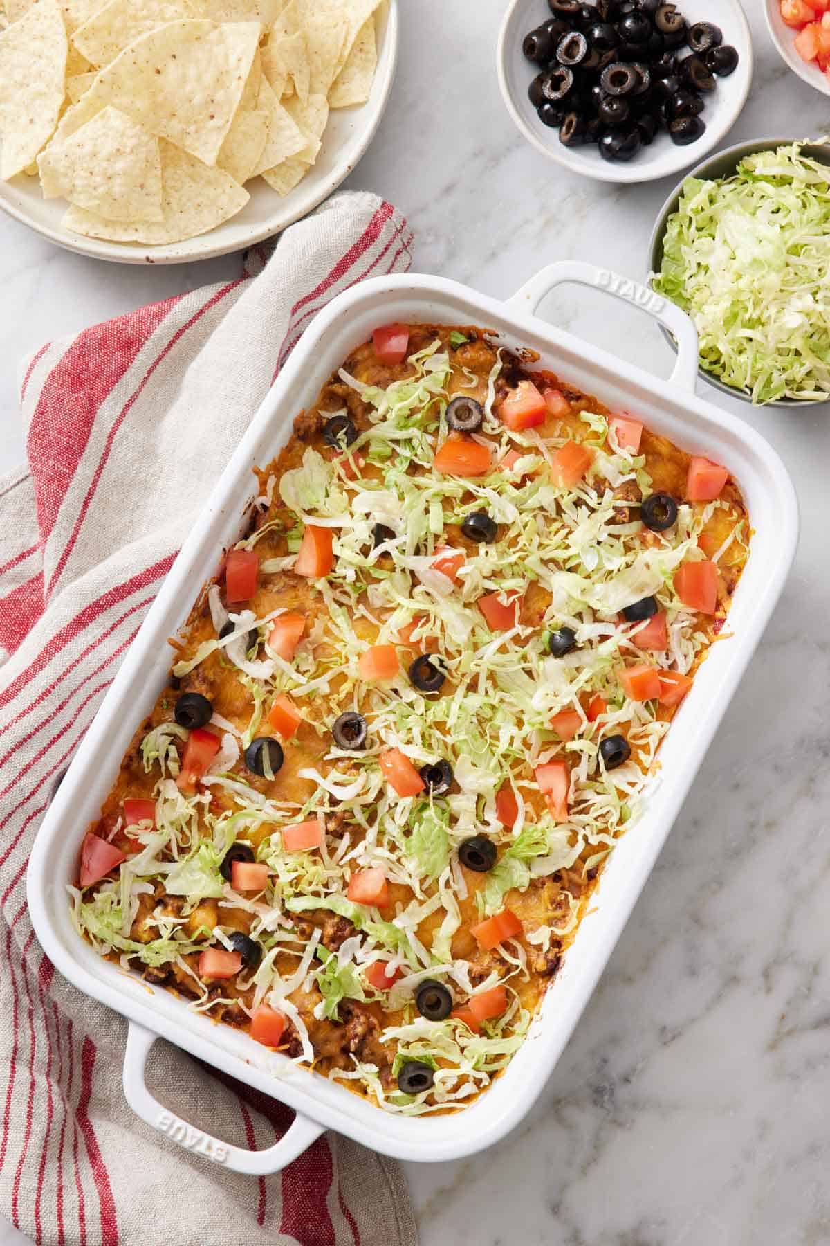 Overhead view of a baking dish with a taco casserole topped with shredded lettuce, diced tomatoes, and olives. More toppings and tortilla chips on the side.