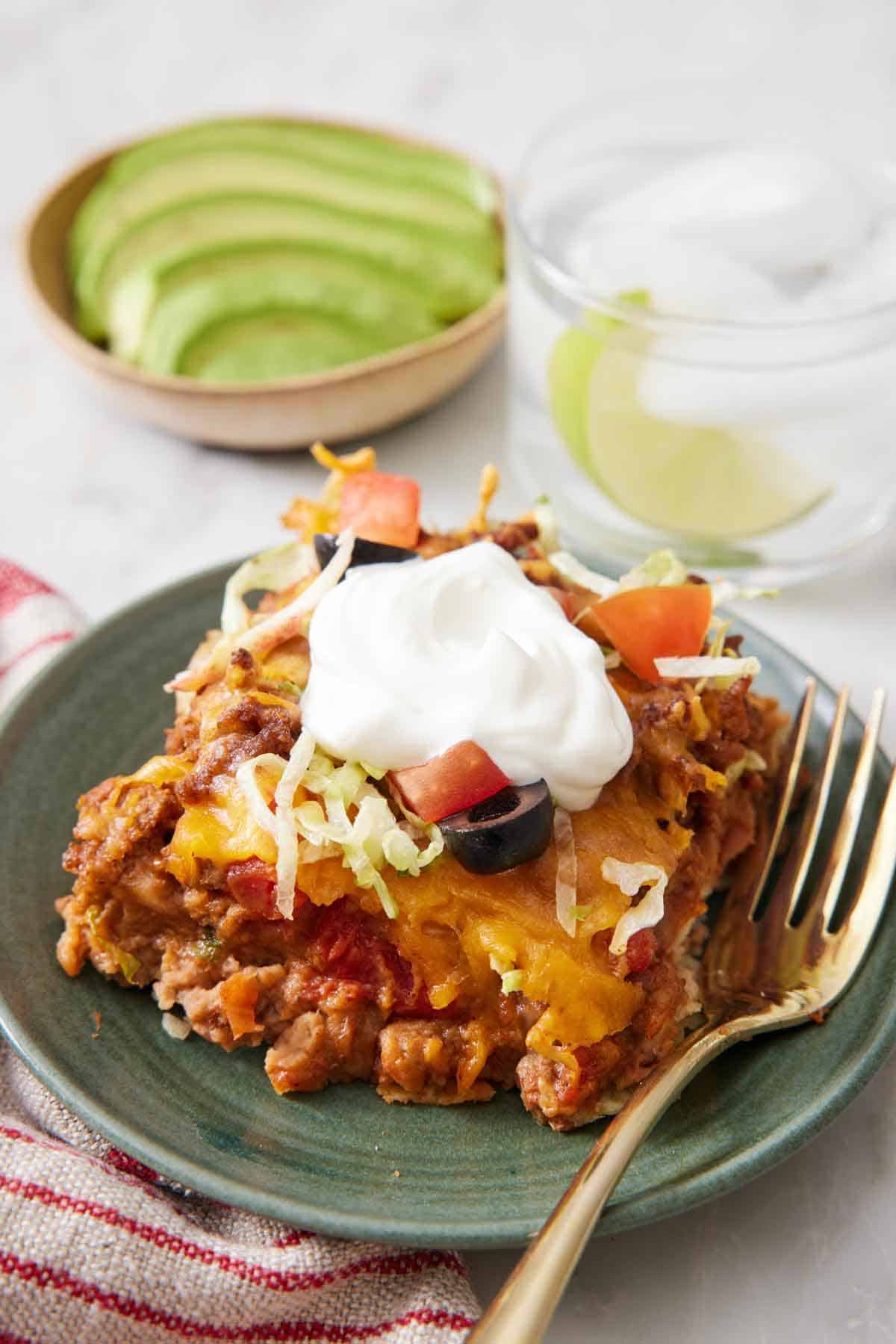 A plate with a serving og taco casserole topped with sour cream. A drink and sliced avocado in a bowl in the background.