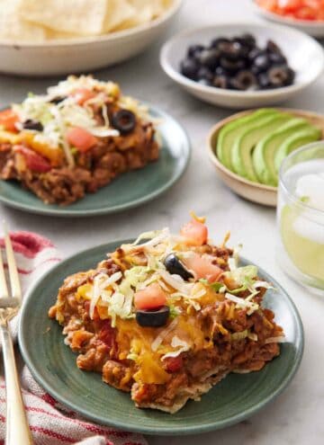 A plate with a serving of taco casserole. Behind it, another plated serving, a drink, a bowl of sliced avocado, a bowl of black olives, and bowl of tortilla chips.