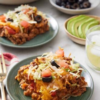 Pinterest graphic of a plate with a serving of taco casserole. In the back is another plated serving, a drink, a bowl of sliced avocado, a bowl of black olives, and bowl of tortilla chips.