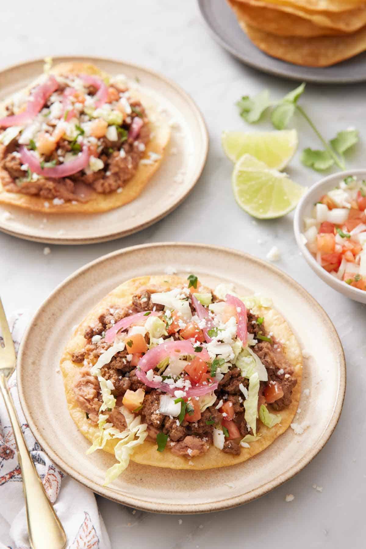 A plate with a tostada with a second plate in the background along with cut limes, cilantro, and a bowl of pico de gallo.