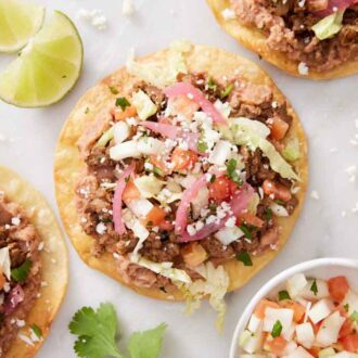 Overhead view of tostadas on a marble surface with crumbled cheese, lime wedges, cilantro, and a bowl of pico de gallo around them.