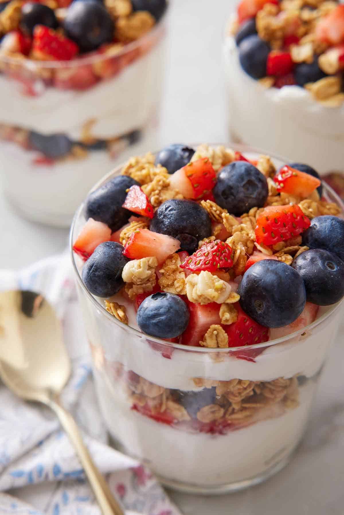 A slightly overhead view of a glass of yogurt parfait with two more in the background and a spoon on the side.