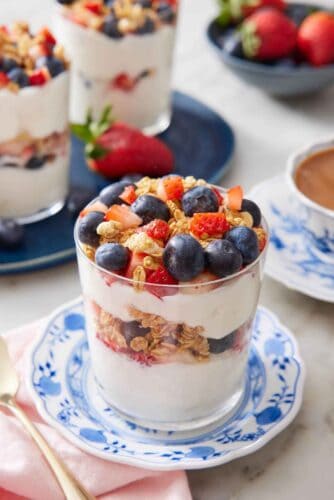 A glass of yogurt parfait on a plate with a platter in the background with two more parfaits.