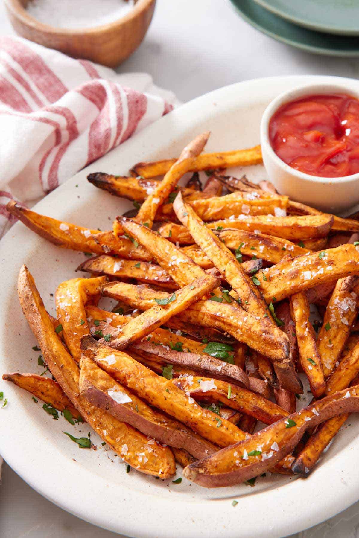 A white platter of air fryer sweet potato fries garnished with parsley and flakey salt with a bowl of ketchup.
