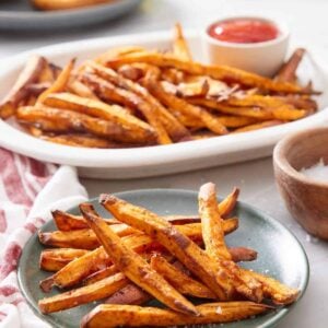 A green plate with air fryer sweet potato fries with a platter more in the background along with ketchup and a burger being assembled.