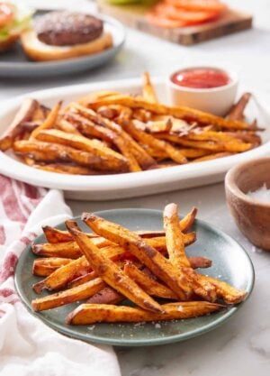 A green plate with air fryer sweet potato fries with a platter more in the background along with ketchup and a burger being assembled.