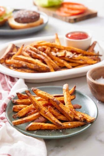 A green plate with air fryer sweet potato fries with a platter more in the background along with ketchup and a burger being assembled.