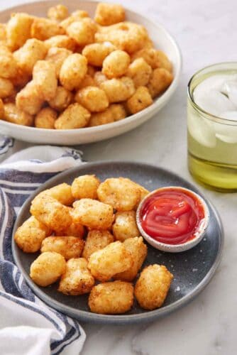 A plate of air fryer tater tots with a bowl of ketchup. A drink in the background along with a large bowl of more tater tots.