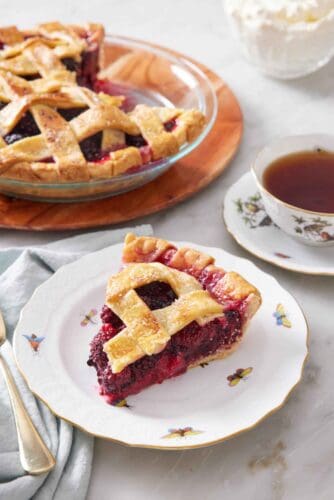 A plate with a slice of blackberry pie with the rest of the pie in the background along with a mug of tea.
