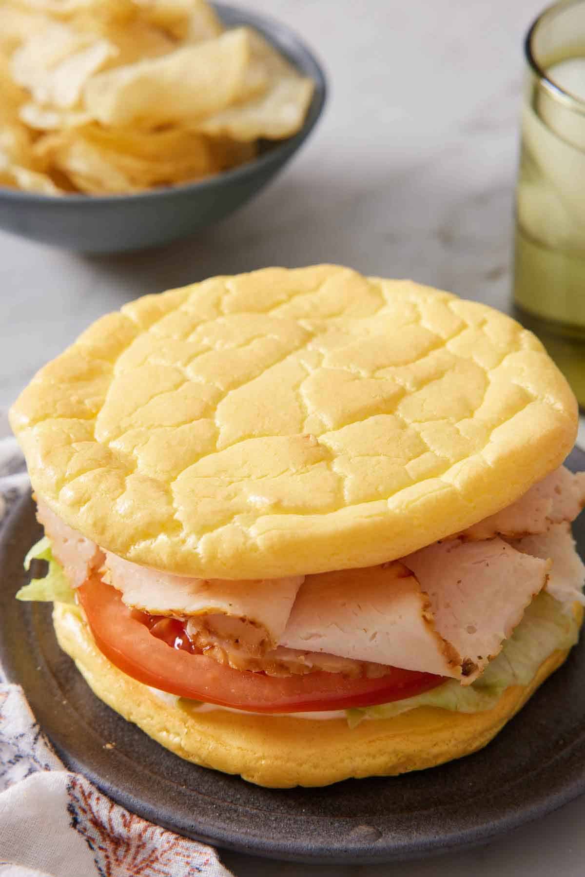 A plate with a sandwich made with cloud bread. A bowl of chips in the background.