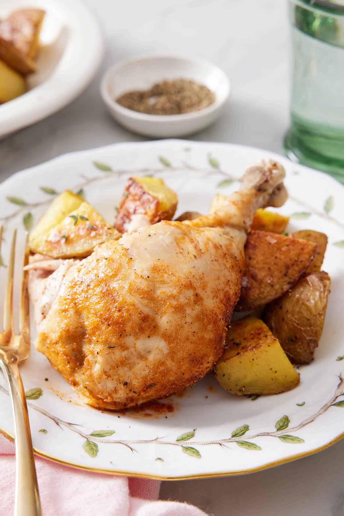 A plate with an instant pot whole chicken leg over roast potatoes. A bowl of pepper in the background along with a glass of water.