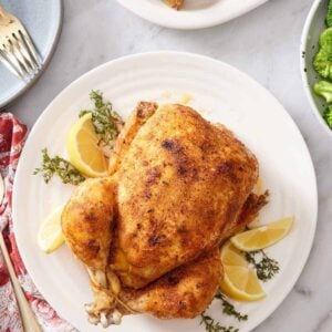 Overhead view of an instant pot whole chicken over fresh thyme and lemon wedges on a white plate. Roast potatoes on the side.