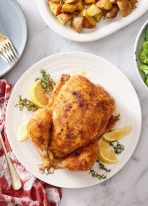 Overhead view of an instant pot whole chicken over fresh thyme and lemon wedges on a white plate. Roast potatoes on the side.