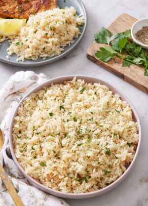 A large plate of rice pilaf with chopped parsley garnish. Another plate in the background with rice pilaf along with a lemon wedge and meat.