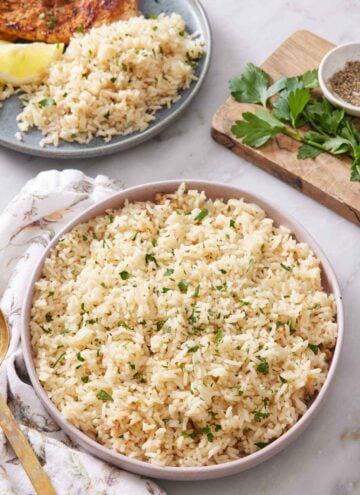 A large plate of rice pilaf with chopped parsley garnish. Another plate in the background with rice pilaf along with a lemon wedge and meat.