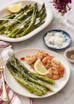 A plate of shrimp with air fryer asparagus and a lemon wedge. A platter of more air fryer asparagus in the back along with a bowl of parmesan.