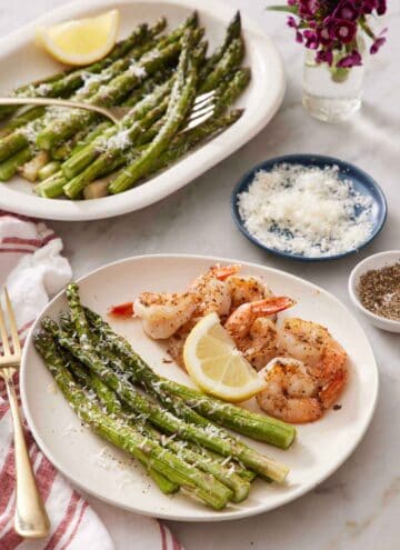 A plate of shrimp with air fryer asparagus and a lemon wedge. A platter of more air fryer asparagus in the back along with a bowl of parmesan.