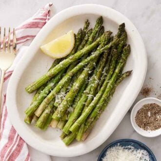Overhead view of air fryer asparagus in a white platter with a lemon wedge. A fork, bowl of pepper, and bowl of parmesan beside it.