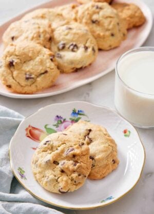 A plate with two air fryer cookies with a glass of milk and a platter of more cookies in the back.