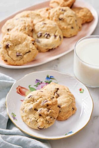 A plate with two air fryer cookies with a glass of milk and a platter of more cookies in the back.