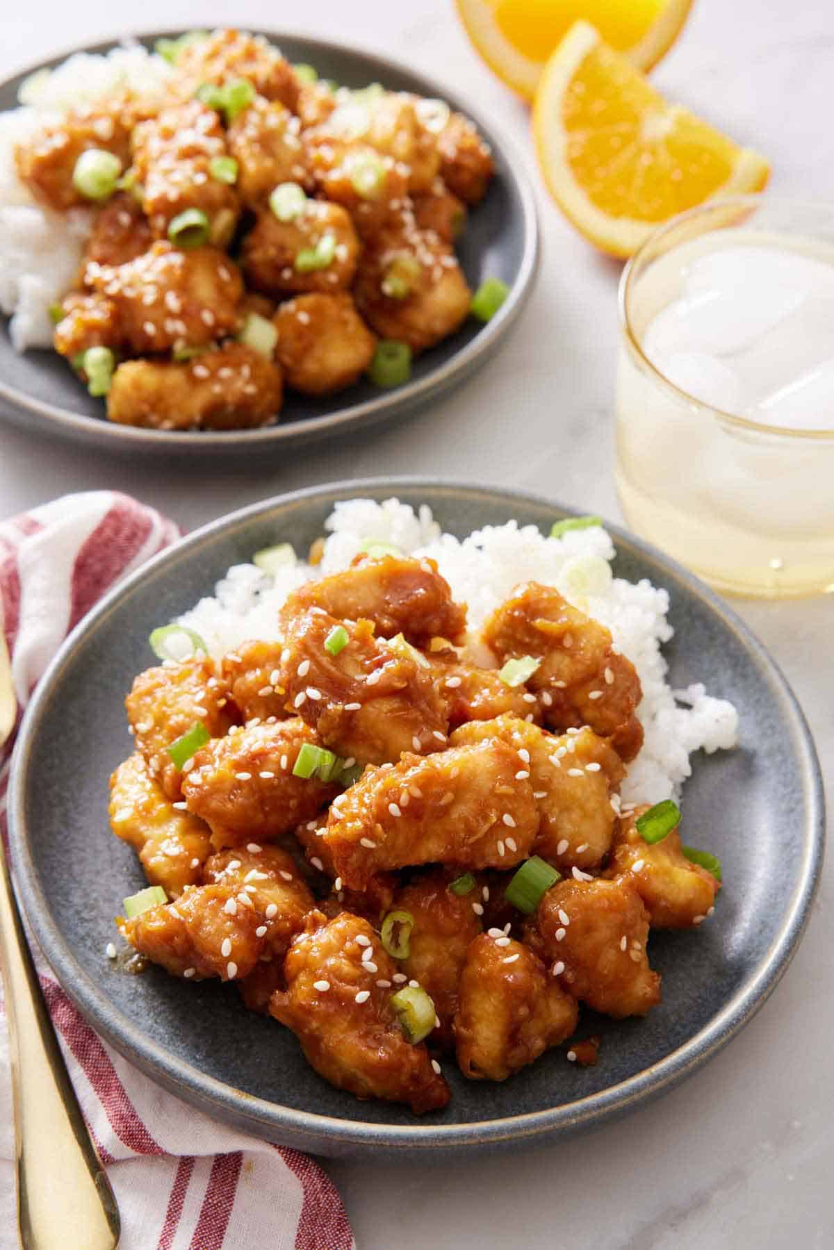 A plate of rice and air fryer orange chicken garnished with sesame seeds and green onions. Another plate in the back along with a drink and orange wedges.