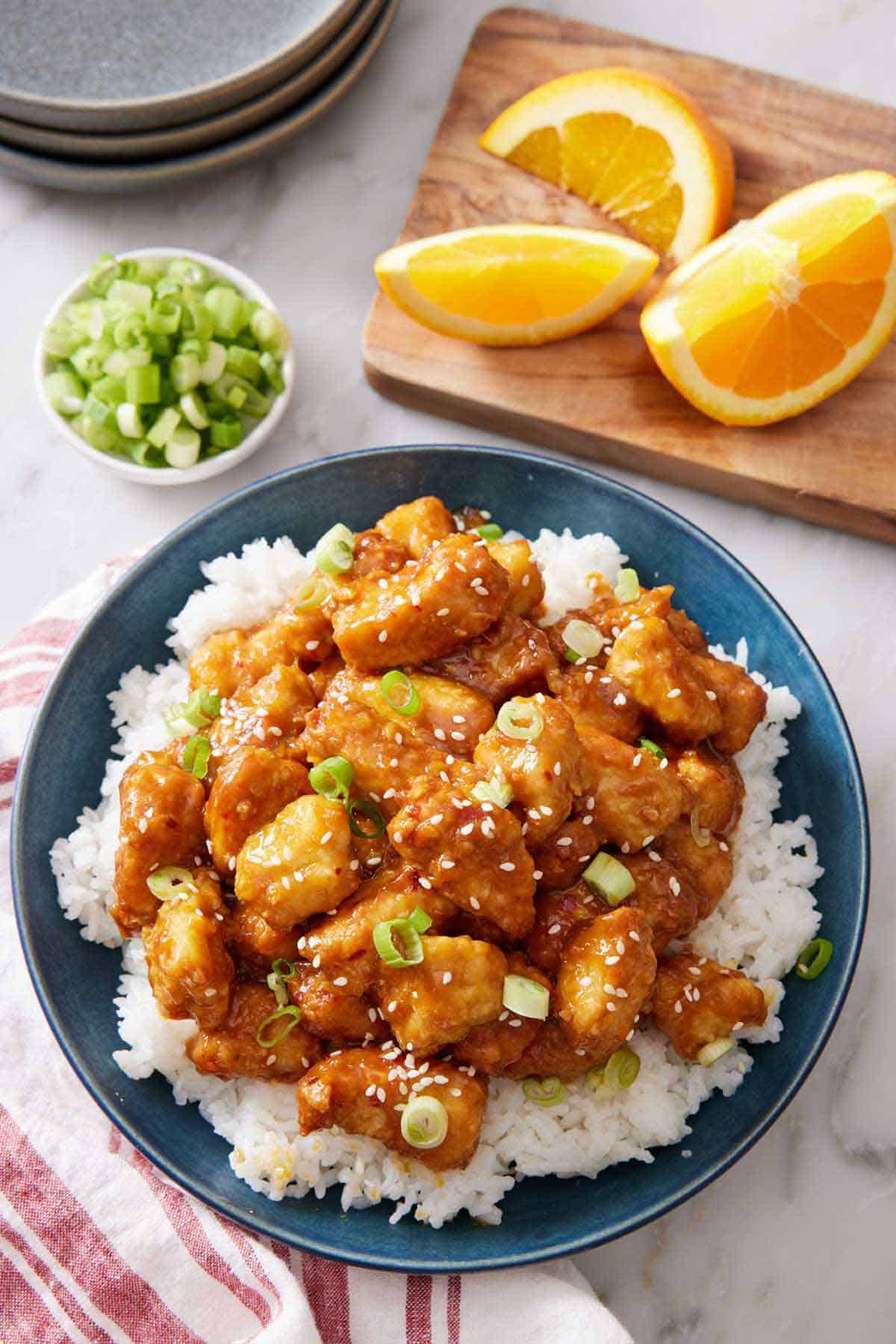 An overhead view of a plate of rice with air fryer orange chicken on top. A board with a cut orange and a bowl of green onions in the back.