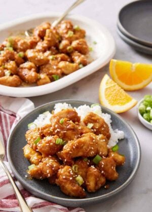 A plate with air fryer orange chicken over rice. A platter in the background with more chicken. Orange wedges on the side.